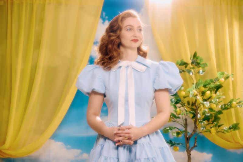 Singer-songwriter Ashley Ann in a pale blue dress with a white bow, standing between yellow curtains beside a lemon tree.