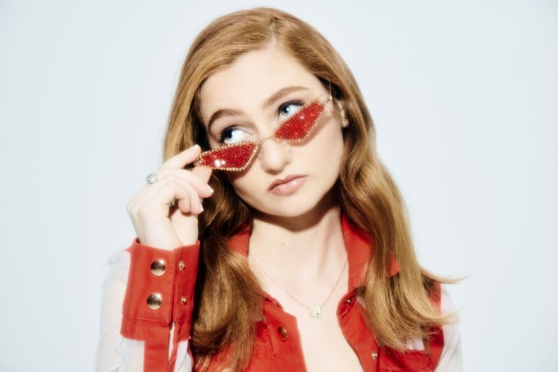 Singer-songwriter Ashley Ann lowering red jeweled cat-eye sunglasses, wearing a red jacket, against a white backdrop.